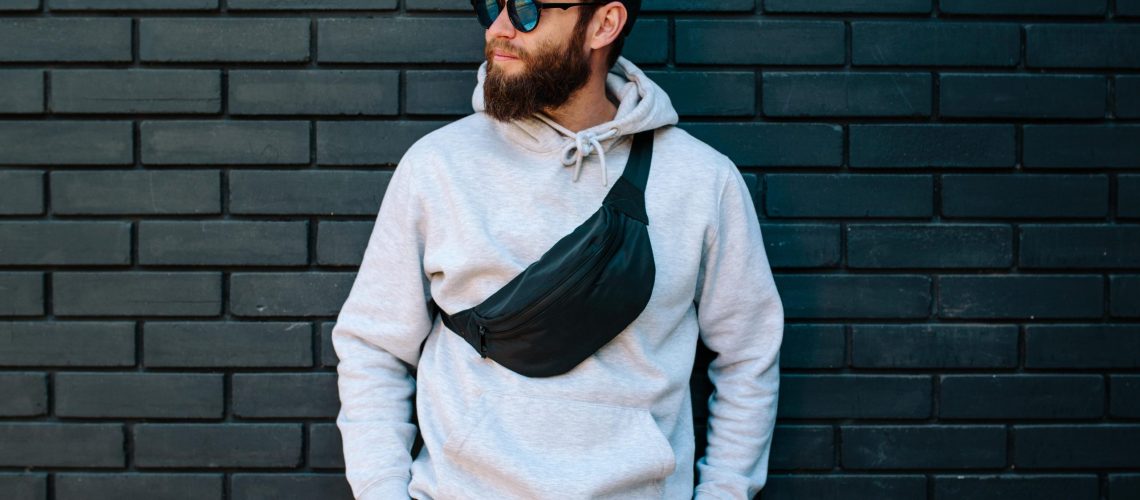 Handsome hipster guy wearing gray blank hoodie, black cap and black waist bag with space for your logo or design. Custom belt bags